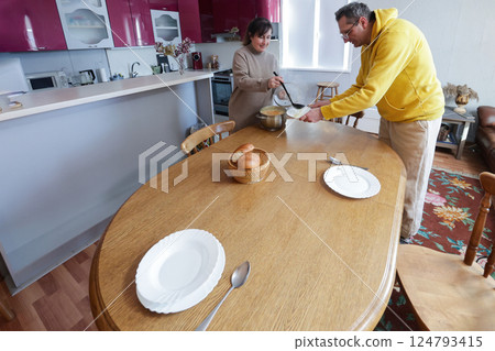 Two people are preparing to eat together as they set table with plates and utensils in cozy kitchen with wooden dining table. 124793415