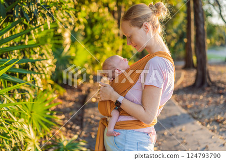 A loving mother walks through a peaceful park, carrying her baby securely in a sling. A heartwarming moment of maternal bonding, babywearing, and the joy of motherhood in nature A loving mother walks through a peaceful park, carrying her baby securely in a sling. A heartwarming moment of maternal bonding, babywearing, and the joy of motherhood in nature 124793790