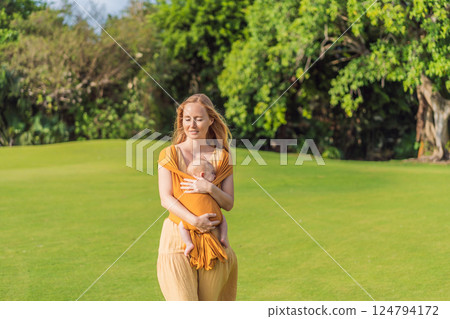 Mother holding her little baby in a yellow sling in the park. Warm and loving family moment. Babywearing, parenting, and mother-child bonding concept 124794172