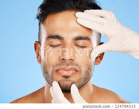 Cosmetic, plastic surgery and face of young man, for skincare and white gloves on blue studio background for dermatology care. Makeup, Indian male and skin rejuvenation for wellness and collagen. Cosmetic, plastic surgery and face of young man, for skincare and white gloves on blue studio background for dermatology care. Makeup, Indian male and skin rejuvenation for wellness and collagen. 124794315