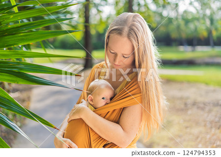 Mother holding her little baby in a yellow sling in the park. Warm and loving family moment. Babywearing, parenting, and mother-child bonding concept Mother holding her little baby in a yellow sling in the park. Warm and loving family moment. Babywearing, parenting, and mother-child bonding concept 124794353