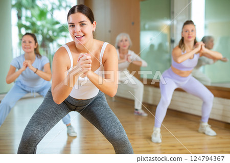 Cheerful young mature female dancers in sportswear dancing with legs wide apart during exercising with coach in gym Cheerful young mature female dancers in sportswear dancing with legs wide apart during exercising with coach in gym 124794367