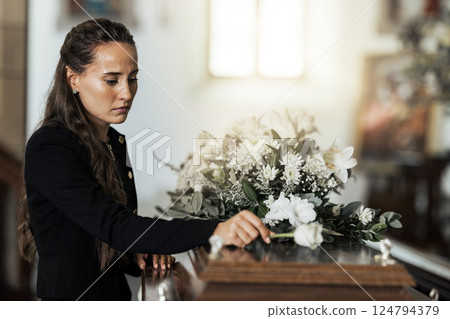 Funeral, sad and woman with flower on coffin after loss of a loved one, family or friend. Grief, death and young female putting a rose on casket in church with sadness, depression and mourning 124794379