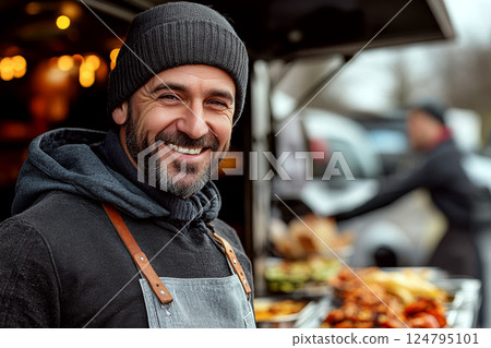 Smiling man wearing a beanie and apron stands outdoors near a food stand with blurred lights in the background. Concept of street food culture. For food vendor photo. 124795101