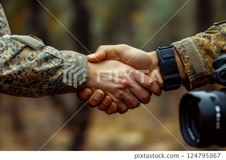 Two people in camouflage uniforms shaking hands outdoors with part of a camera visible. Concept of solidarity and partnership. For unity and teamwork. Two people in camouflage uniforms shaking hands outdoors with part of a camera visible. Concept of solidarity and partnership. For unity and teamwork. 124795867