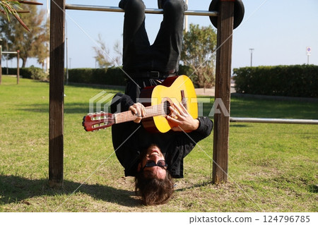 Young Man Plays Guitar Music Hanging Upside Down In An Outdoor Park Young Man Plays Guitar Music Hanging Upside Down In An Outdoor Park 124796785