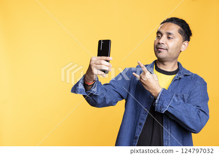 Excited confident man holding his phone to take photos in the studio, feeling pleased with his look and outfit against orange background. Trendy joyous indian person with a positive attitude. Excited confident man holding his phone to take photos in the studio, feeling pleased with his look and outfit against orange background. Trendy joyous indian person with a positive attitude. 124797032