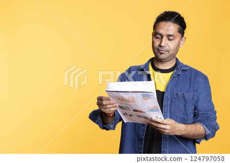Indian accountant doing profit analysis for company and browsing files, standing against orange background. Worker doing financial budgeting and forecasting, looking over paperwork graphs. 124797058