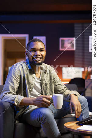 African american smiling man sipping on coffee working remotely, finishing his tasks at the coffee table in the living room. Casual relaxed person browsing the internet and multitasking at home. 124797073