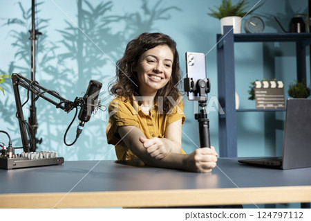 Expressive cheerful content creator using smartphone on selfie stick to shoot video. Joyous influencer filming vlog with mobile phone in studio, using professional microphone Expressive cheerful content creator using smartphone on selfie stick to shoot video. Joyous influencer filming vlog with mobile phone in studio, using professional microphone 124797112