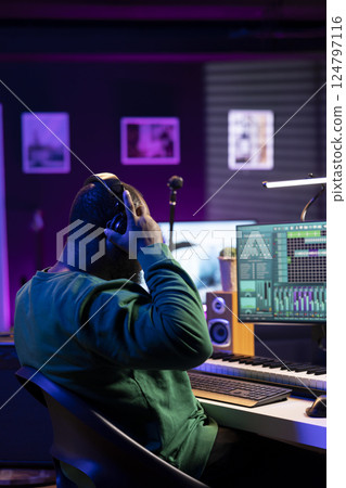 Musician playing piano keys and editing to improve the sound quality of audio recordings in his home studio, putting on headset. Music producer uses midi controller to manipulate sounds. 124797116