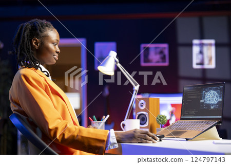 Smiling woman using laptop in apartment, updating artificial intelligence machine learning algorithms, writing code. Upbeat teleworking IT expert working from home uses programming to upgrade AI 124797143