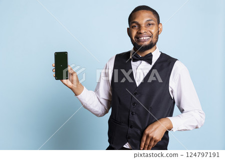 Hotel worker bellhop points at phone with isolated mockup display, showing green screen against blue background in studio. Hotelier doorman greeting resort guests at reception. Hotel worker bellhop points at phone with isolated mockup display, showing green screen against blue background in studio. Hotelier doorman greeting resort guests at reception. 124797191