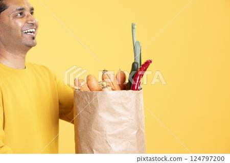 Happy man walking with paper bag in hands filled with vegetables, coming from supermarket, studio background. Sustainable living person holding groceries shopping bag after doing purchases in store 124797200