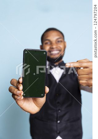 African american bellhop presenting green screen on a phone, posing with hospitality industry suit and tie in studio. Young hotelier personnel creates an isolated advertisement. African american bellhop presenting green screen on a phone, posing with hospitality industry suit and tie in studio. Young hotelier personnel creates an isolated advertisement. 124797204