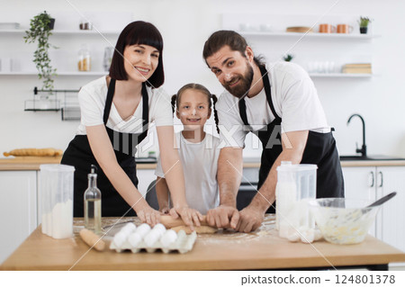 Caucasian family of parents and a young daughter cooking in modern kitchen, bonding, enjoying preparing and rolling dough together, showcasing teamwork, happiness, and quality time in a pleasant 124801378