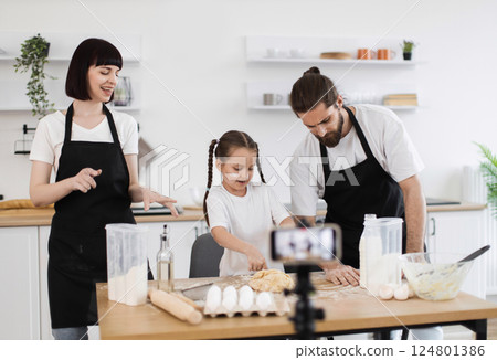 Caucasian family cooking together in kitchen, recording video while bonding over food preparation. Parents and child in aprons kneading dough, smiling and enjoying shared cooking experience. 124801386