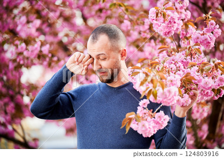 Man allergic suffering from seasonal allergy at spring in blossoming garden at springtime. Bearded man feeling itchy eyes in front of blooming tree. Spring allergy concept. 124803916