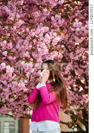 Woman allergic suffering from seasonal allergy at spring in blossoming garden at springtime. Woman sneezing and blowing nose using nasal handkerchief in front of blooming tree. Spring allergy concept. 124803917