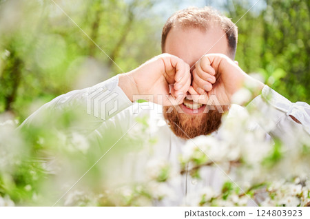 Man allergic suffering from seasonal allergy at spring in blossoming garden at springtime. Close up of bearded man sneezing and feeling itchy eyes in front of blooming tree. Spring allergy concept. 124803923