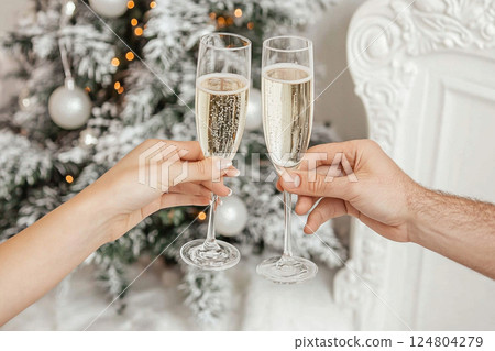 Two hands hold champagne flutes to celebrate a special occasion in a cozy, warmly decorated space featuring a festive Christmas tree adorned with ornaments and lights. 124804279