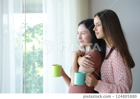 Happy interracial lesbian couple drinking looking through a window Happy interracial lesbian couple drinking looking through a window 124805880