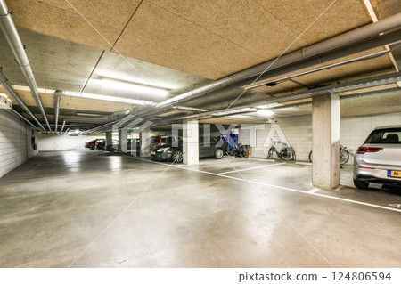 A spacious underground parking lot featuring concrete walls, overhead pipes, and parked vehicles, illuminated by bright ceiling lights. Ideal for urban environments. A spacious underground parking lot featuring concrete walls, overhead pipes, and parked vehicles, illuminated by bright ceiling lights. Ideal for urban environments. 124806594