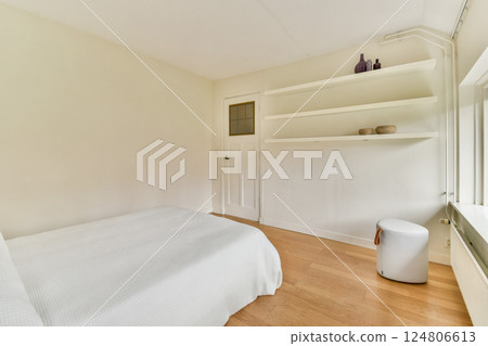 A bright, minimalist bedroom featuring a bed with a white cover, wooden flooring, and simple shelves that enhance the tranquil atmosphere. 124806613