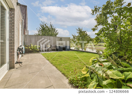 A well-maintained garden featuring lush green grass, plants, and patio furniture under a bright blue sky, perfect for relaxation and outdoor gatherings. 124806666