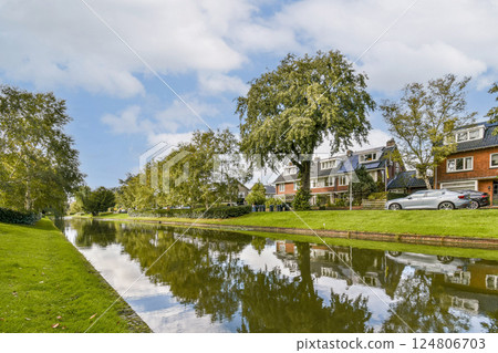 A serene riverside view featuring trees lining a calm waterway, complemented by charming nearby houses. The clear sky enhances the picturesque landscape. A serene riverside view featuring trees lining a calm waterway, complemented by charming nearby houses. The clear sky enhances the picturesque landscape. 124806703