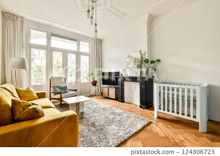 A modern living room featuring a cozy ambiance, yellow sofa, light colors, wooden flooring, and a baby crib, ideal for family living spaces. A modern living room featuring a cozy ambiance, yellow sofa, light colors, wooden flooring, and a baby crib, ideal for family living spaces. 124806723