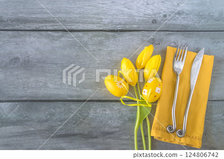 Place setting for easter dinner or lunch: bouquet of yellow tlips, easter eggs and fork and knife on a yellow napkin, wooden background; copy space 124806742