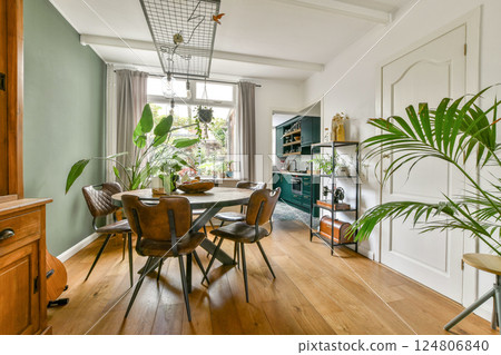 A stylish modern kitchen featuring a wooden dining table surrounded by chairs, vibrant plants, and soft natural light streaming through large windows. A stylish modern kitchen featuring a wooden dining table surrounded by chairs, vibrant plants, and soft natural light streaming through large windows. 124806840