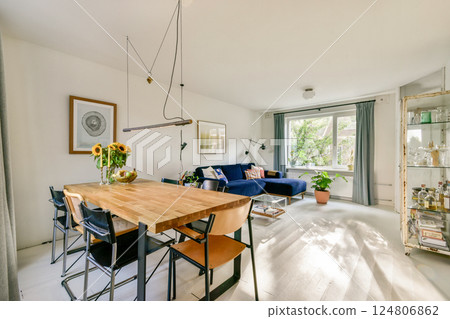 A stylish living space featuring a wooden dining table, blue sofa, and large windows bringing in natural light and greenery. 124806862
