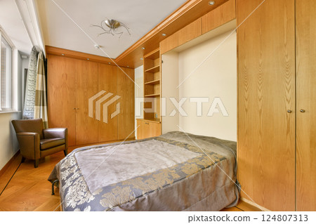 An elegantly designed bedroom featuring wooden cabinetry, a comfortable bed, and a stylish chair, showcasing modern interior decor and lighting. 124807313