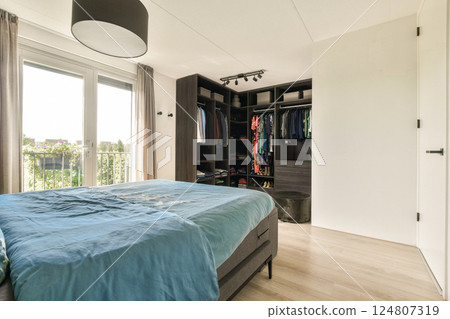 A spacious modern bedroom featuring a neatly organized closet, large windows allowing natural light, and stylish furnishings that enhance comfort and aesthetics. 124807319