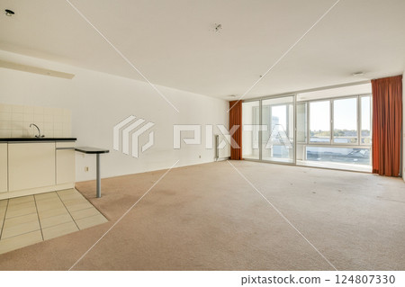 Spacious living room features large windows, beige carpet, and a minimalist kitchen setup, providing a bright and airy atmosphere. 124807330