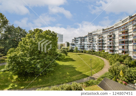 A serene view of a green park with a lush tree, juxtaposed against contemporary residential buildings under a bright sky. A serene view of a green park with a lush tree, juxtaposed against contemporary residential buildings under a bright sky. 124807451