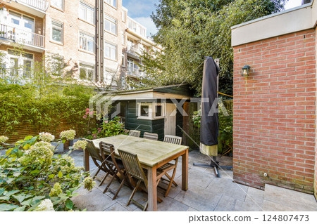 A serene garden with a wooden table and chairs surrounded by greenery and a small shed. Perfect for relaxing or social gatherings. 124807473