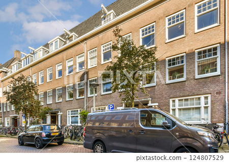 A picturesque urban street lined with parked vehicles and bicycles, surrounded by charming brick buildings and trees under a clear blue sky. A picturesque urban street lined with parked vehicles and bicycles, surrounded by charming brick buildings and trees under a clear blue sky. 124807529