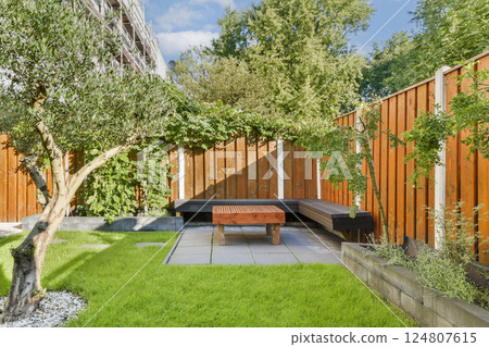 A peaceful outdoor space featuring a vibrant green lawn, a wooden bench, and a beautiful tree, all enclosed by stylish wooden fencing. 124807615