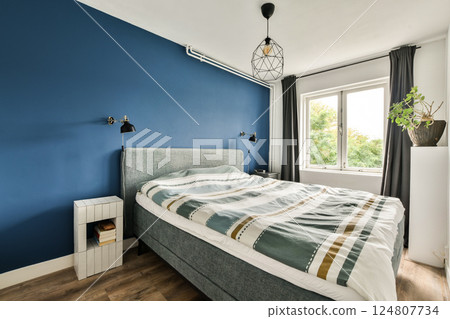 A modern bedroom featuring a blue accent wall, large windows, and a stylish bedspread, creating a serene and inviting atmosphere. A modern bedroom featuring a blue accent wall, large windows, and a stylish bedspread, creating a serene and inviting atmosphere. 124807734