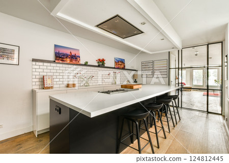 A sleek and stylish modern kitchen featuring a white countertop, black bar stools, and elegant decor. The space is bright and inviting, perfect for cooking and entertaining. 124812445