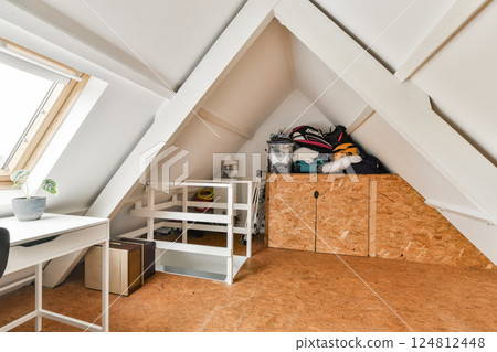 A modern attic workspace includes cozy decor, a desk, and organized storage. The room is brightened by natural light from a large window. 124812448