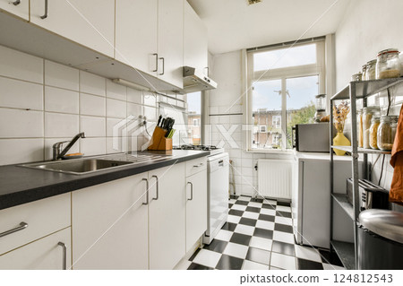 A sleek modern kitchen featuring a minimalist design with white cabinetry, black and white tiled flooring, and ample natural light streaming through the window. 124812543