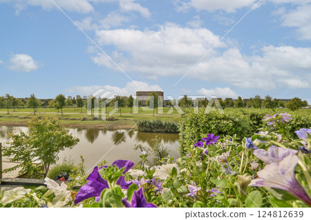 A picturesque view featuring vibrant flowers in the foreground, a tranquil pond, lush greenery, and a cloudy sky, creating a peaceful natural scene. A picturesque view featuring vibrant flowers in the foreground, a tranquil pond, lush greenery, and a cloudy sky, creating a peaceful natural scene. 124812639