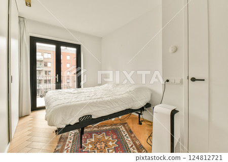 A spacious and bright bedroom featuring a white bed, wooden floors, and large windows allowing natural light. The room has a clean and modern aesthetic. 124812721
