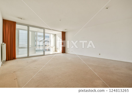 A spacious, empty living room featuring large glass windows that allow natural light. The room has light-colored walls and carpet, offering a minimalist aesthetic. A spacious, empty living room featuring large glass windows that allow natural light. The room has light-colored walls and carpet, offering a minimalist aesthetic. 124813265
