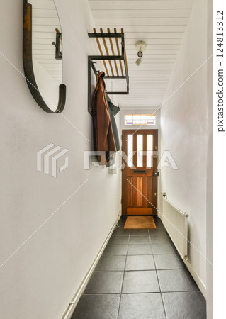 A bright and sleek hallway featuring a wooden door, stylish mirror, and organized coat rack, creating an inviting entryway in a contemporary home. 124813312