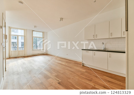 A spacious, minimalist design featuring a bright room with natural light, hardwood flooring, and a sleek kitchen area with cabinetry. 124813329